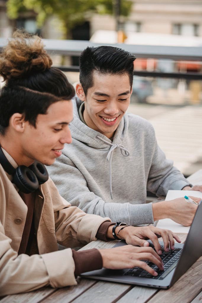 Two young men working together on a laptop outside, demonstrating teamwork and collaboration.